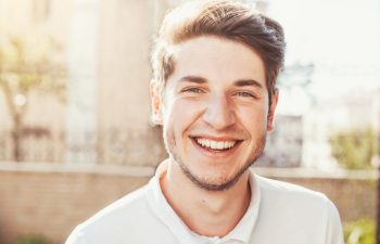 A smiling person with short hair and a light beard standing outdoors, wearing a white shirt. The background is softly blurred, suggesting a sunny day.